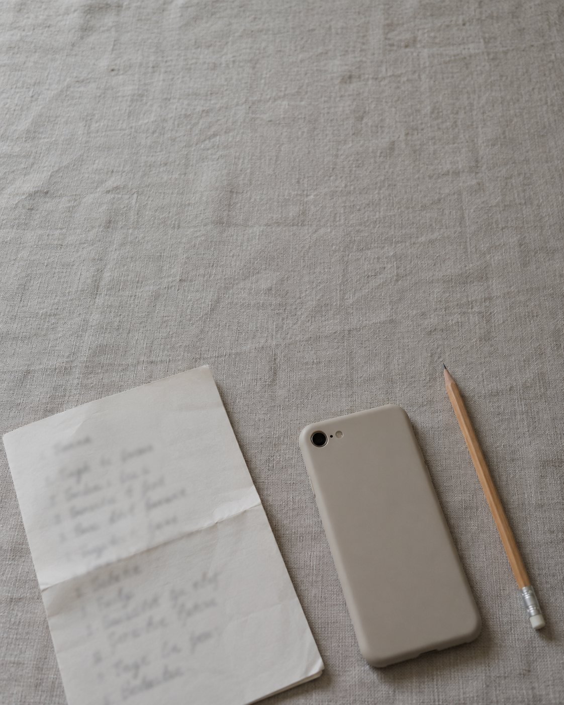 A handwritten list, a phone face down and a pencil on grey linen
